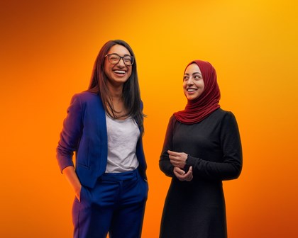Two women laughing together in front of an orange background